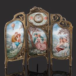 RELOJ DE SOBREMESA ART NOUVEAU Tríptico decorado con esmaltes de escenas campestres y marco con detalles Art Nouveau en latón. La hoja central lleva embutido un reloj con esfera de porcelana blanca y numeración arábiga. Sistema de carga cuerda-llave. En e